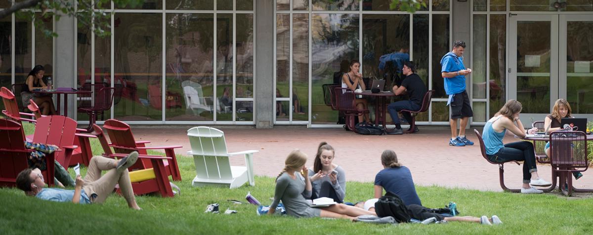 group of students lounging on grass and sitting at tables outside glass-walled building
